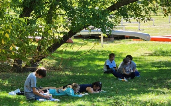 Six students write while sitting and laying on the grass under a tree with canoes in the background.
