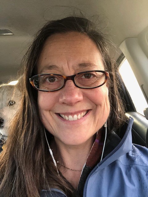 Selfie of woman with long brown hair and glasses sitting in car with her dog visible behind her.