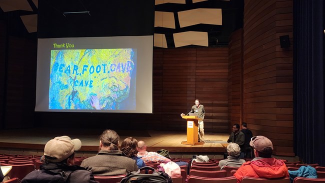 Presenter at podium with slide titled The Bear Foot Cave