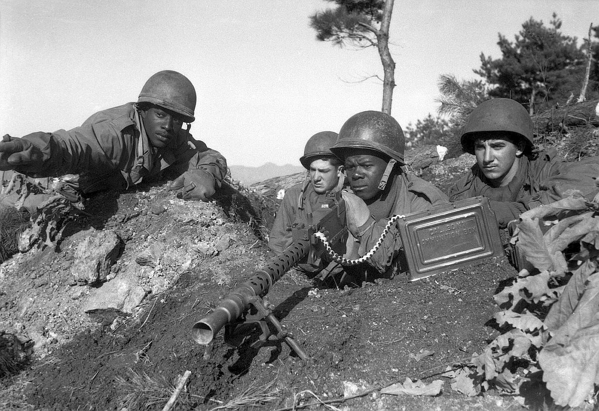 Black and white photo of 4 American soldiers in a fox hole manning a machine gun. 2 soldiers are Black and 2 soldiers are White.