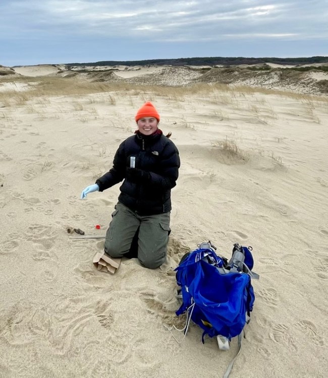 Scientist sits on beach dune with backpack in front of her smiling.