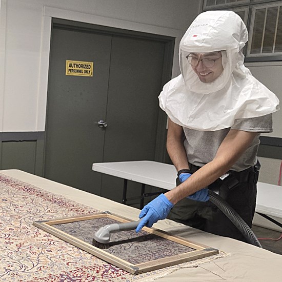 A park employee in protective clothing vacuums a colorful rug.