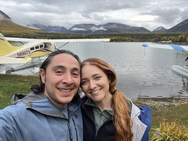 Two people stand in front of a lake with a yellow float plane to the left in the water. In the background are mountains with overcast conditions