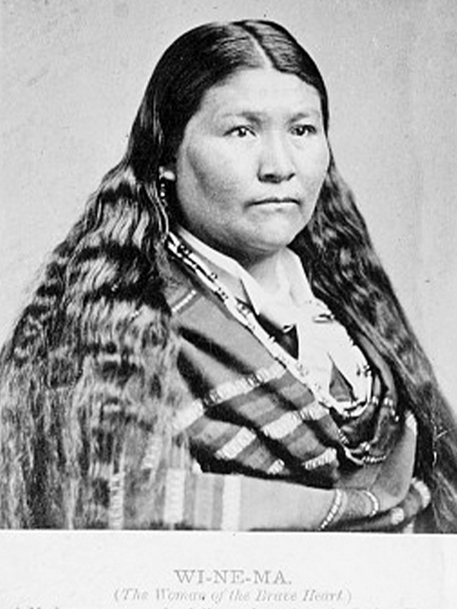 Native American woman with long hair, wrapped in stripped shawl and beaded necklace, gazes at camera.