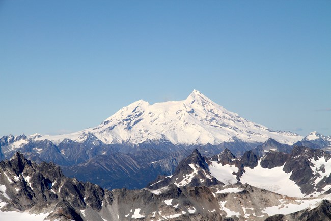 Image of blue skies with snow capped stratovolcano and jagged peaks in foreground.
