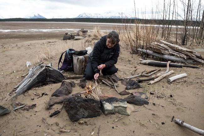 Image of a woman making a fire in a fire pit, along a lakeshore.
