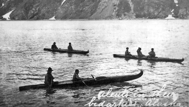 3 baidarkas, or kayaks, on the water, with its occupants waving at the camera.