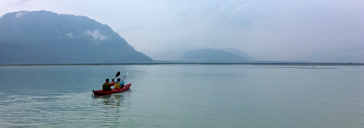 Two persons in a kayak paddling out in a bay with mountains in the far distance