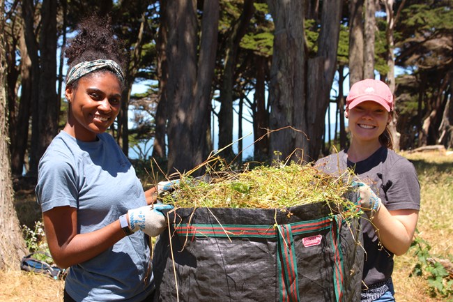 Two women hold up a huge bag overflowing with freshly pulled weeds.