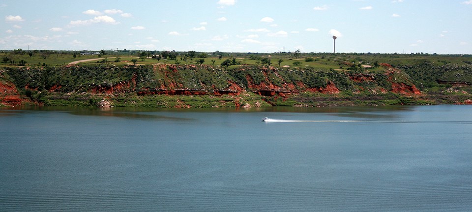 Lake Meredith A boat speeds across a large lake bounded by reddish cliffs covered in green plants below a grassy savanna