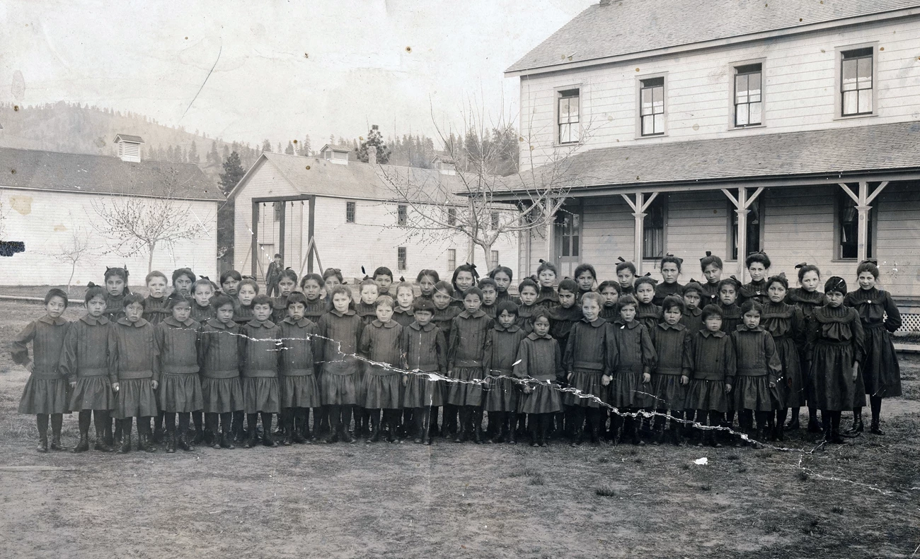 Group of school girls, many Indigenous, at Fort Spokane. Taken c. 1903 Black and white photograph of several rows of identically dressed Indigenous students standing in front of buildings