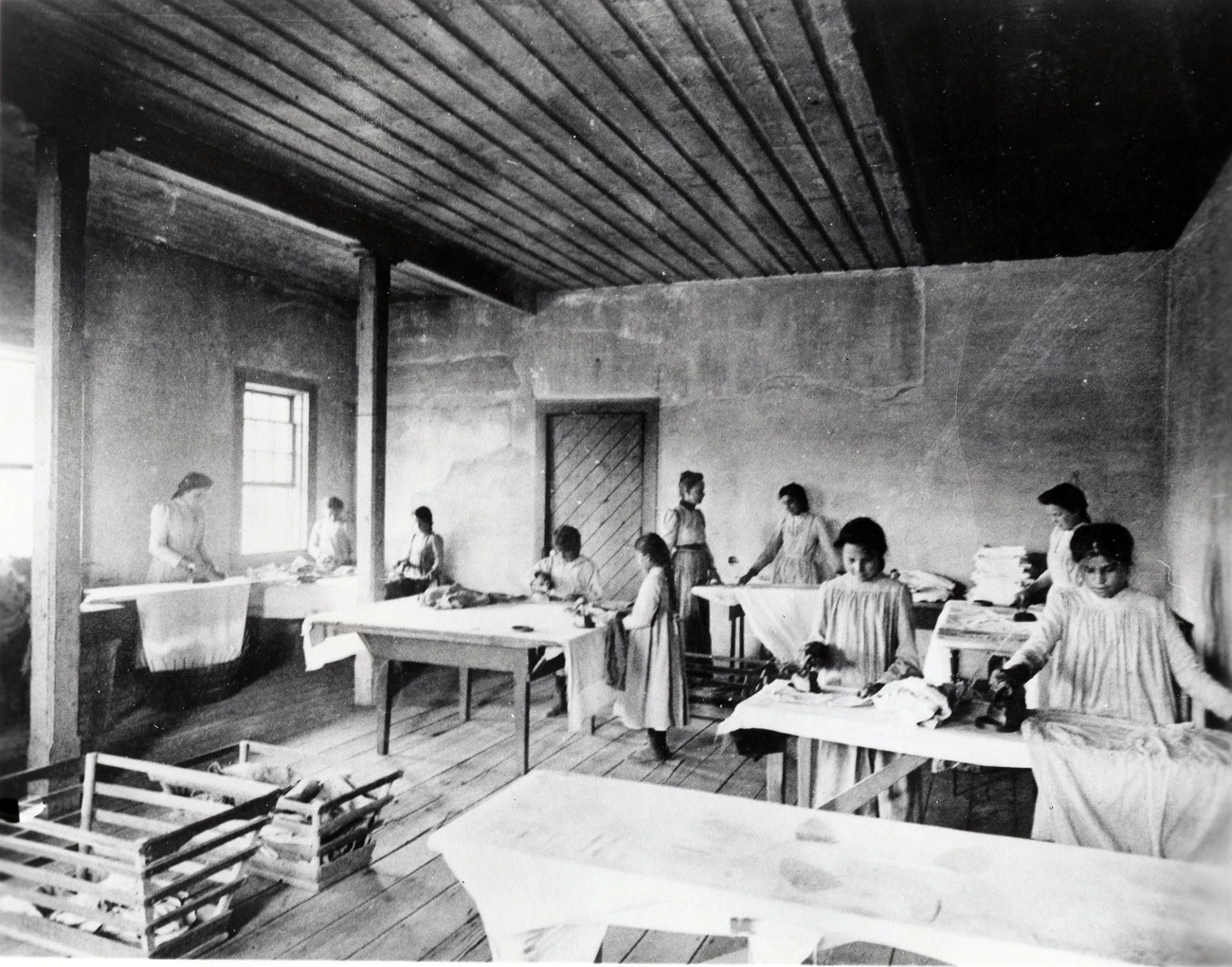 The photographer is unknown, but it was likely taken by Frank Fuller Avery, the superintendent of Fort Spokane Indian boarding school. Nine Indigenous girls in western-style dresses and haircuts iron laundry as white teacher looks on.