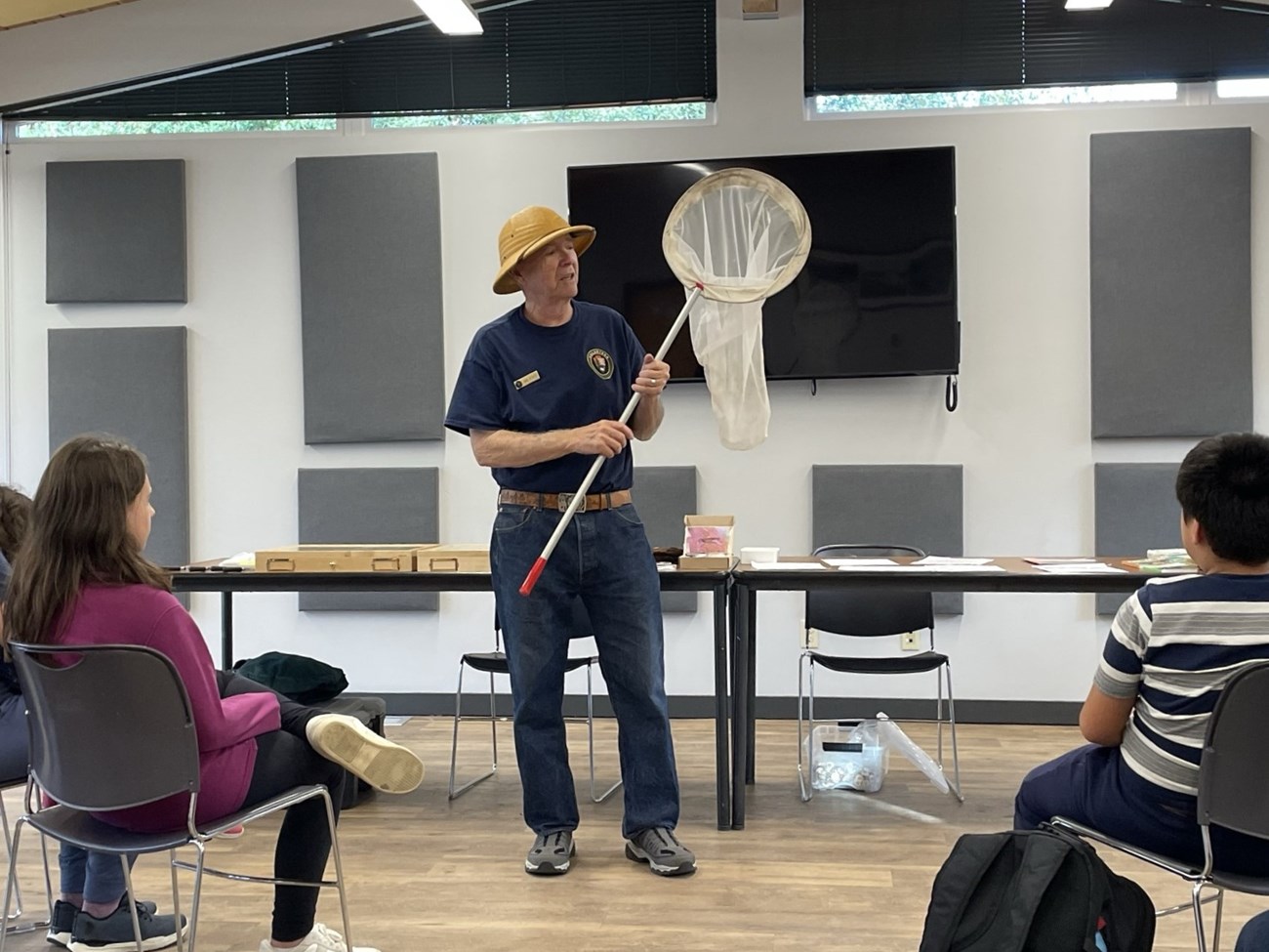 VIP Insect Program at Library Man in a blue shirt and tan field hat holds a large bug net up to an audience.