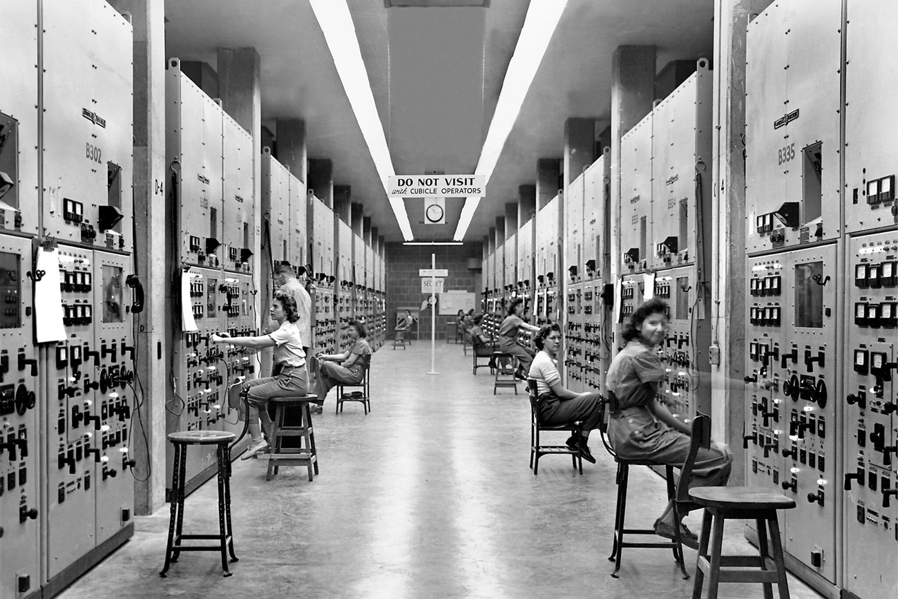 Black and white photo of several women seated along a long hallway covered in knobs and dials.