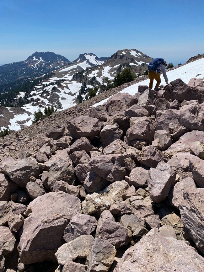 Climbing up to a whitebark pine monitoring plot. Person with backpack climbs up rocky talus on a mountainside with snowfield in background.