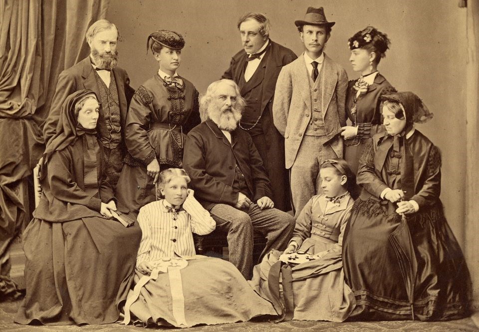 lt= Black and white photograph of ten people standing and seated in studio in Victorian clothing