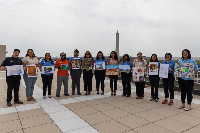 13 individuals hold up art designs with the Washington Monument in the background