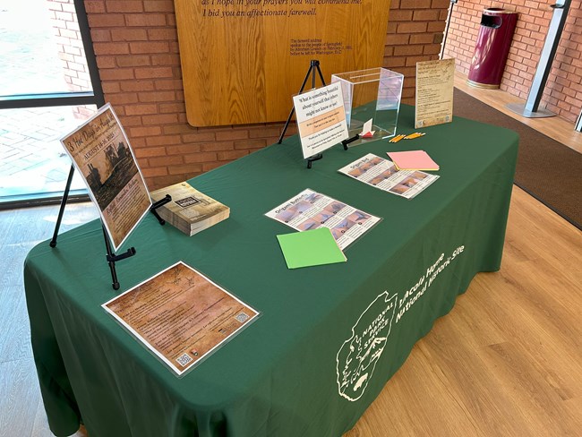 Pieces of origami paper and folding directions lie on a green tabletop inside a room with wood floors and brick walls.