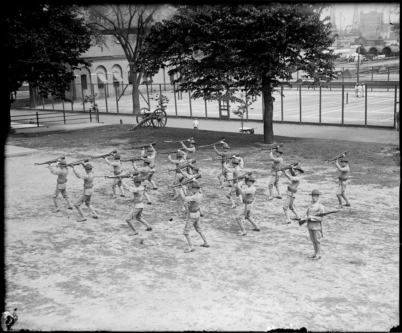 Grid of 16 Marines on a parade ground. They hold bayonets over their heads as an officer watches.