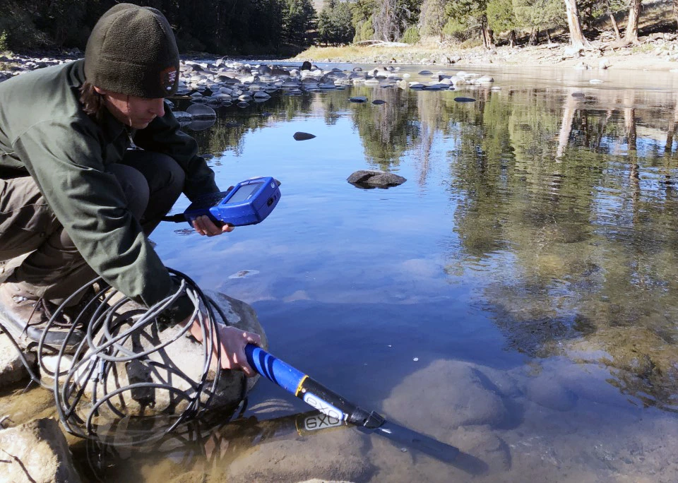 Sampling the Lamar River A scientist crouched down at a river's edge holding a probe in the water and an electronic display in their other hand.