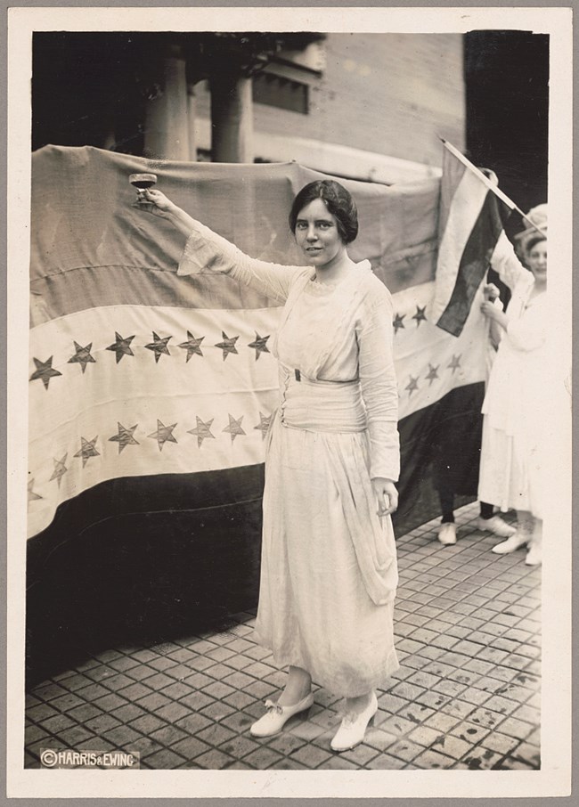 Alice Paul raises a glass in front of a ratification flag with sewn stars