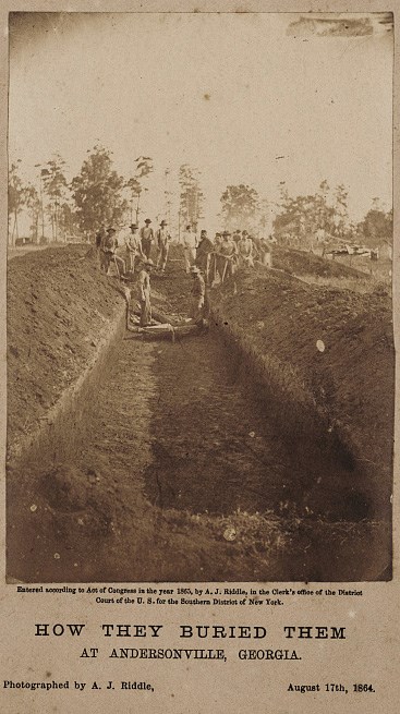 Black and white photograph looking down a deep trench with men in background burying dead prisoners into the trench.
