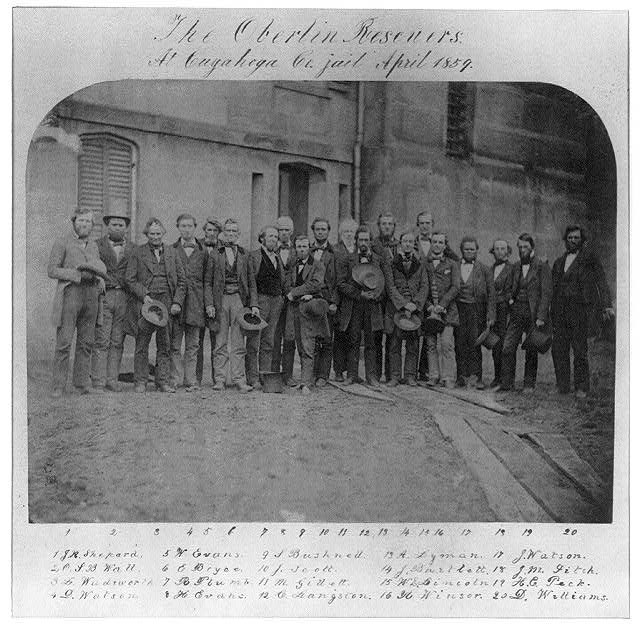 A photograph taken outside depicting 20 men. Above the photograph is cursive writing that reads "The Oberlin Rescuers. At Cuyahoga Co. Jail." Underneath the photograph in cursive writing is the first initial and last name of those depicted.