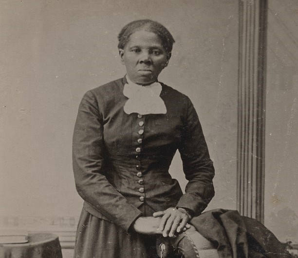 Harriet Tubman as a young woman stands next to a chair, placing her left hand on it. She stares into the camera with a determined facial expression.