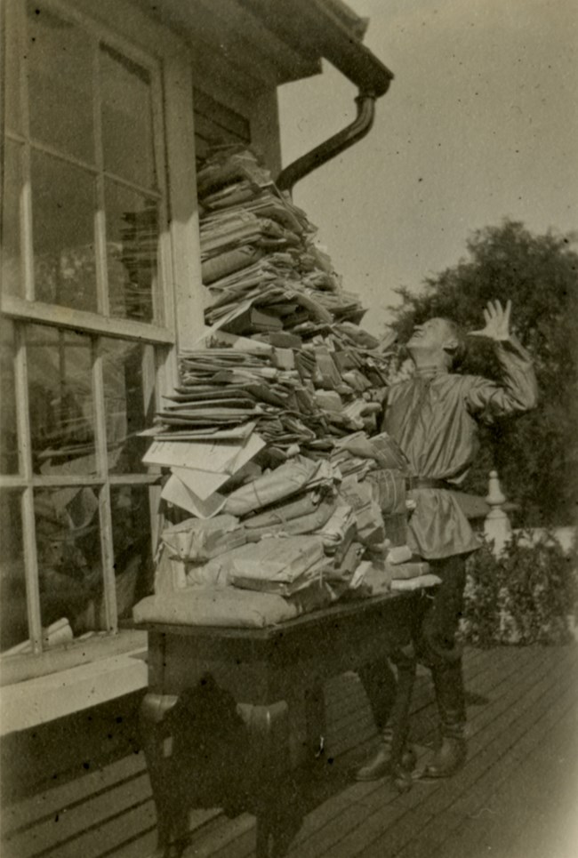 LONG 1002-9-4-1131 White man wearing a Cossack shirt and tall boots poses with his hands in the air looking up at a pile of papers on a table outside.