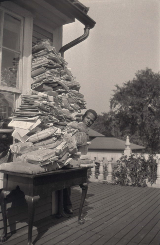 LONG 1002-9-4-1234 Table piled with documents and packages higher than a person's height, with only the head of a Black man visible peeking around the stack.