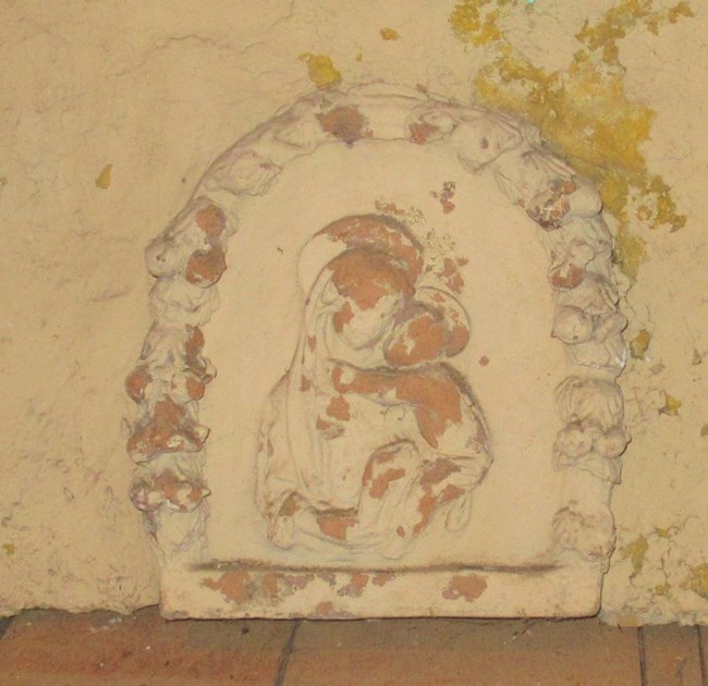 Tan bas relief of woman holding baby in center of arched laurel chain. Paint flaking on surface.