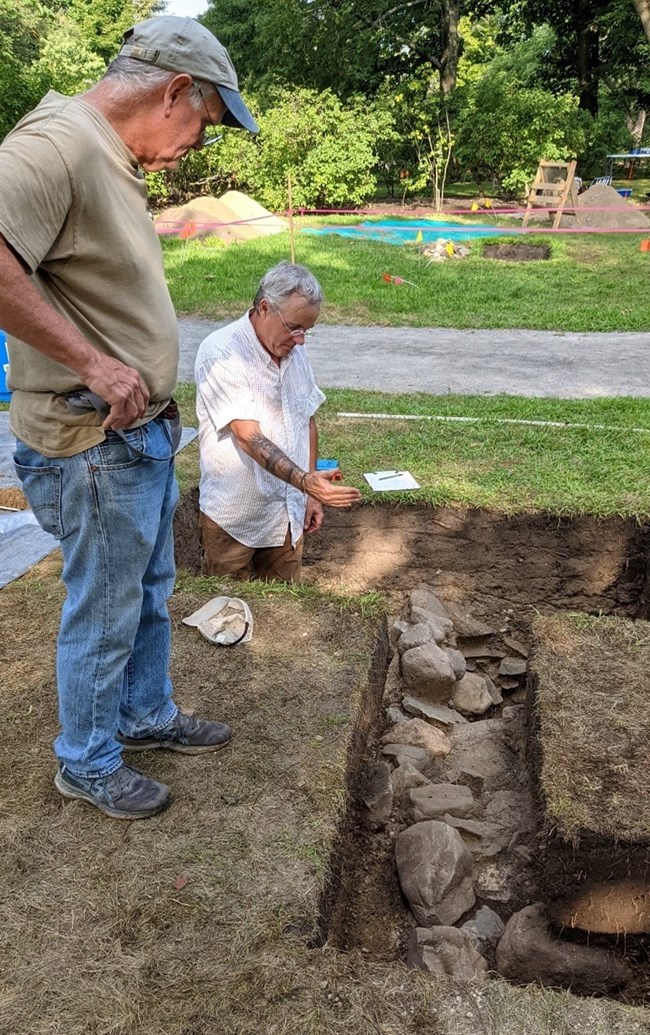 Two men stand in and above an archeology unit with a rock wall at the bottom