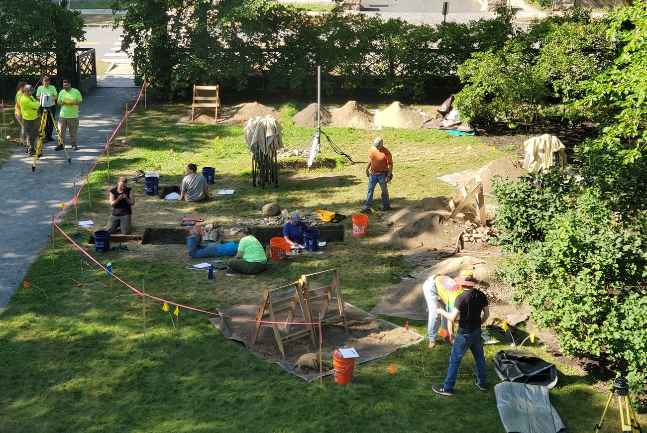 Marrett Cellar Excavation, 2022 11 people stand in and around rectangular holes in a lawn with piles of dirt