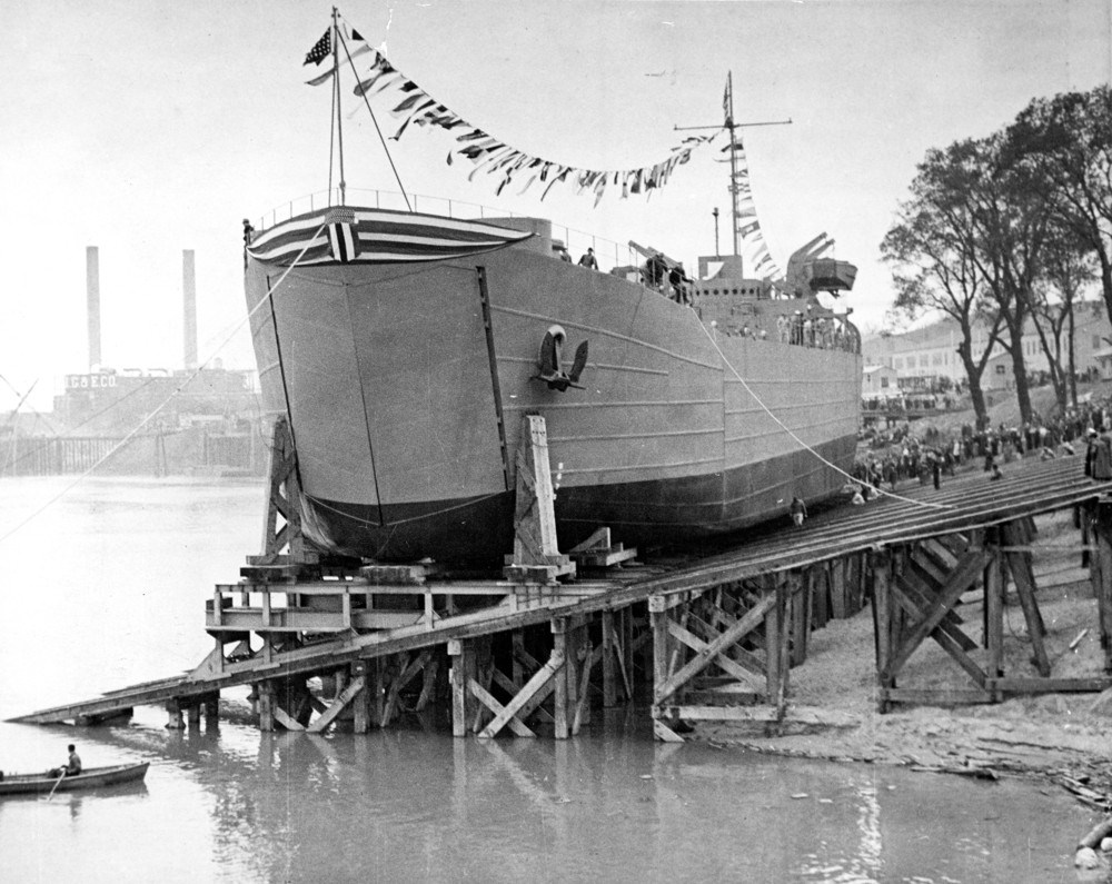 LST 157 in Evansville Ohio. Courtesy of the University of Southern Indiana. Black and white photo of a ship sitting on a wooden scaffolded dock. Flags fly between the mast and smokestacks are visible inbackground.