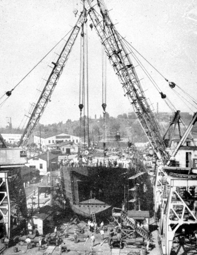 Bird’s eye view of tall cranes and scaffolding supporting an incomplete ship’s hull. Workers move equipment and ladders around the base of the hull. Several workers are also visible at the deck-level.