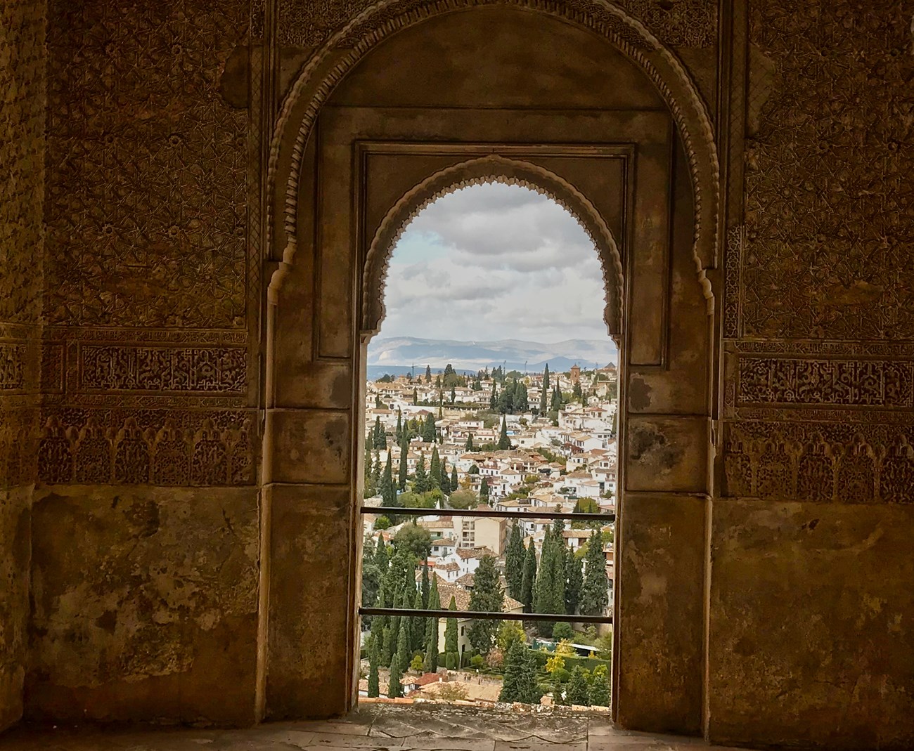 View of Spanish town outside of an ornate window with decorative walls. The town has houses with orange roofs and white stucco and trees are interspersed. Mountains are present in the horizon.