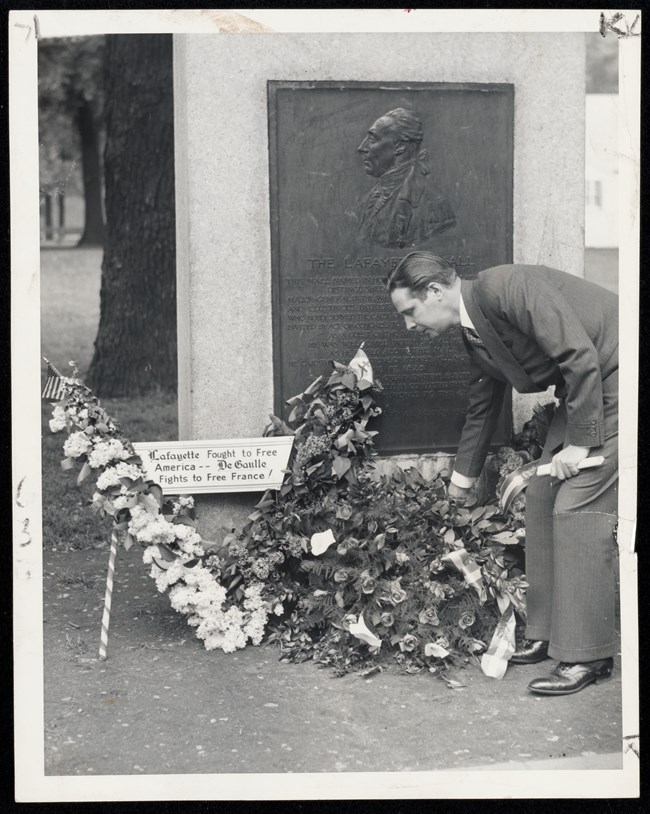 Man lays a wreath and flowers at the base of a stone and metal tablet featuring