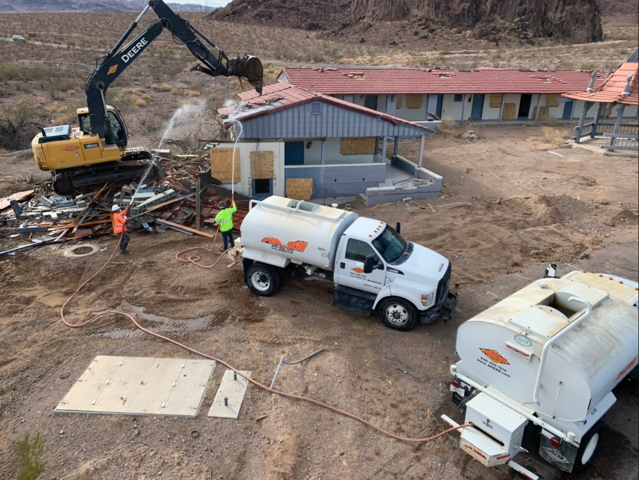 Lake Mead Demolition A yellow construction vehicle tears down an old building. Two construction workers spray water on the demolished area. White trucks appear in the foreground.