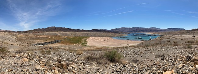 A rocky desert landscape sits in front of a sliver of lake. Blue skies and a mountain range sit in the background