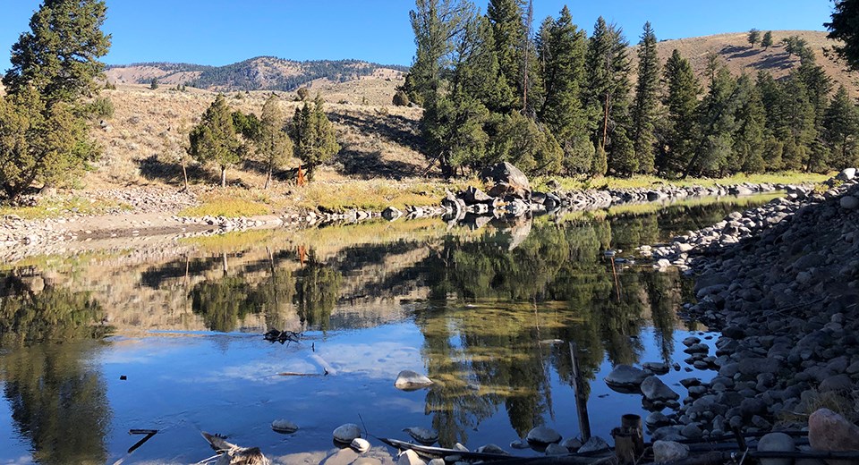 A calm river lined by rocks reflecting the trees and hills behind it.