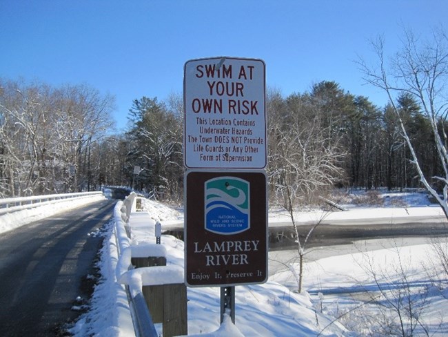 A sign that says "Swim at Your Own Risk" and below a sign that has the wild and scenic river logo and says "Lamprey River"