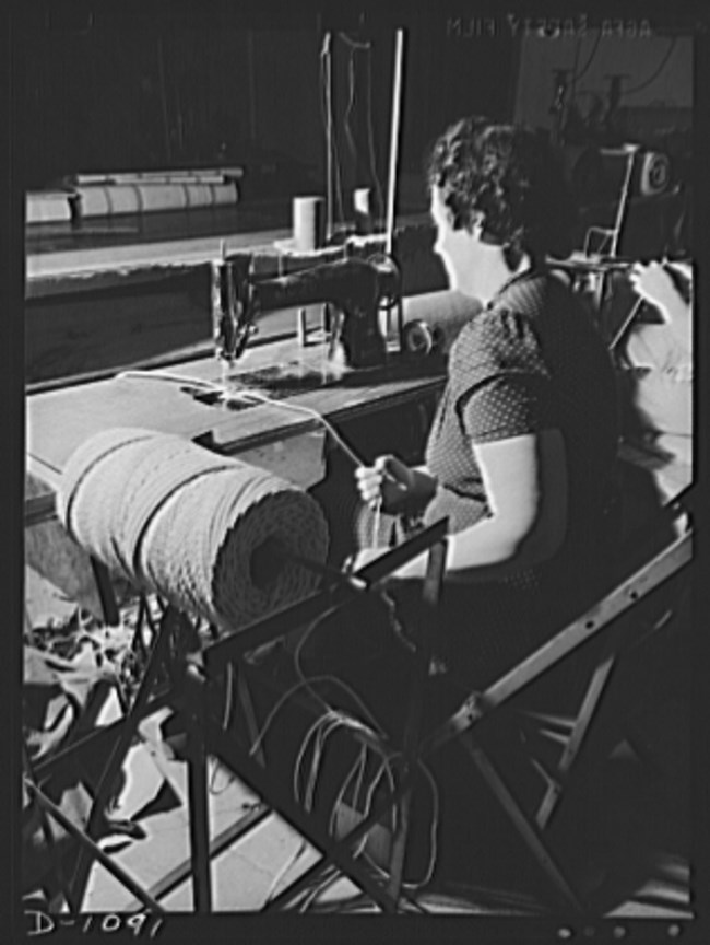Photo 4: Langdon Tent and Awning Co 2 Black and white photo of a white woman with her back to the camera at a sewing machine. A spool of thread in the foreground.