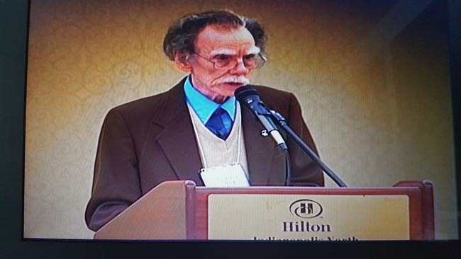 A middle aged man stands behind a podium presenting a lecture.