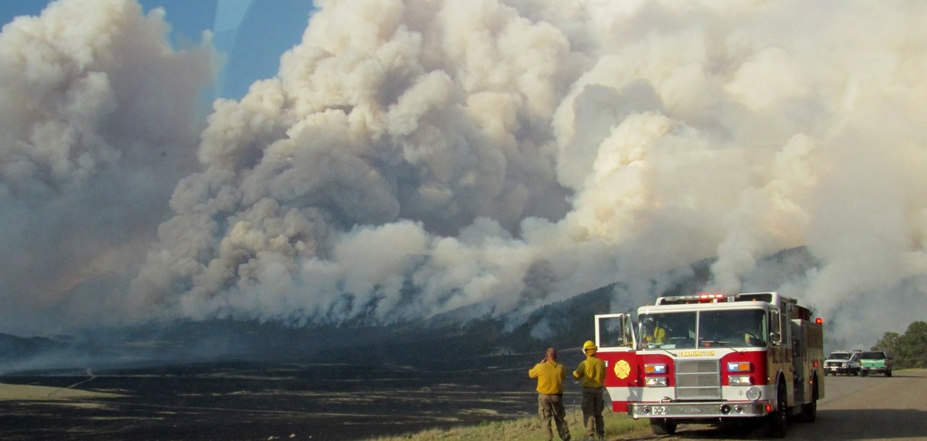 Las Conchas Fire, Valle Grande - photo by J. Dewar Firefighters stand outside of a red fire engine and watch a large wildfire burning in the distance.