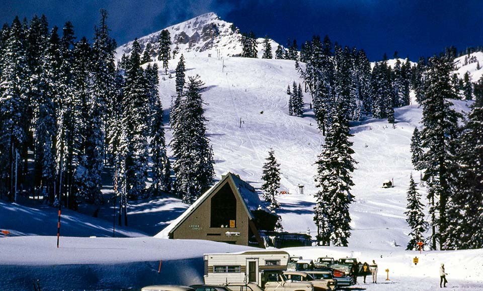 Vehicles parked in front of a chalet backed by a ski hill with a single lift.