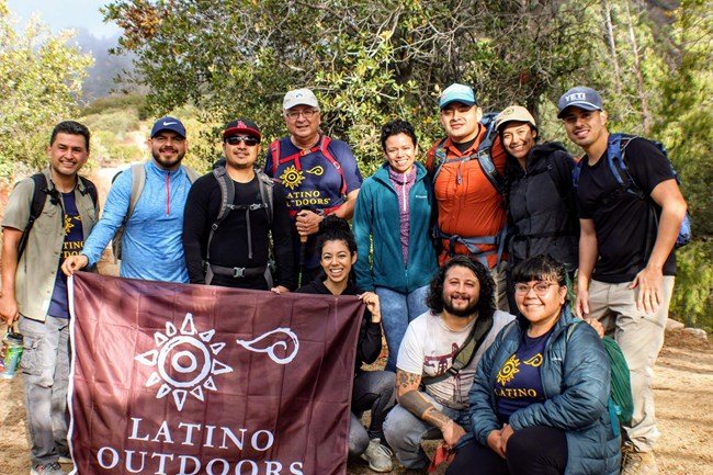 lt= A group of hikers pose for a picture holding a burgundy flag with the Latino Outdoors Logo.