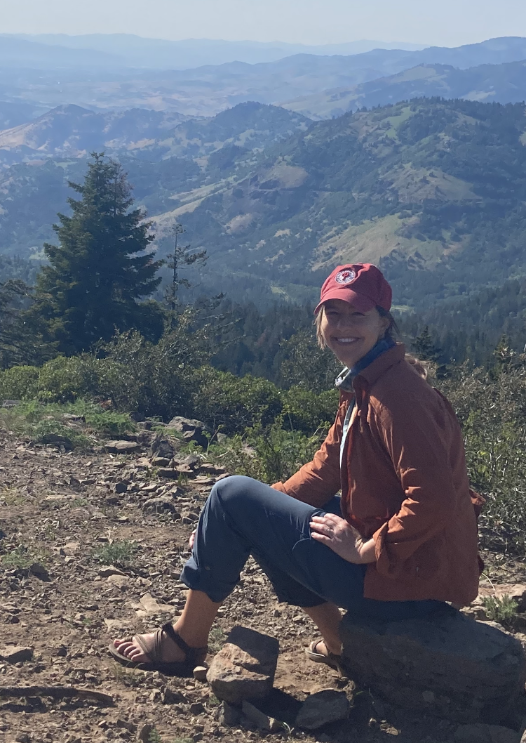 Woman sitting on rock with forested mountains in background.