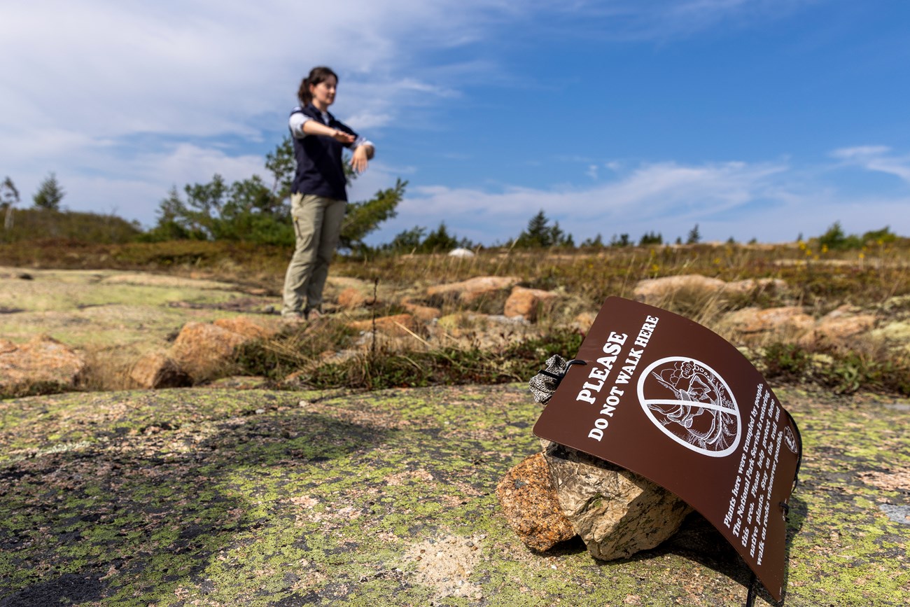 A young woman stands in the background outdoors, with a sign saying “please do not walk here” in the foreground.