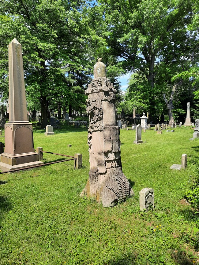 Tall gravestone, shaped like a tree trunk, with an acorn shaped top, in a cemetery, surrounded by other gravestones