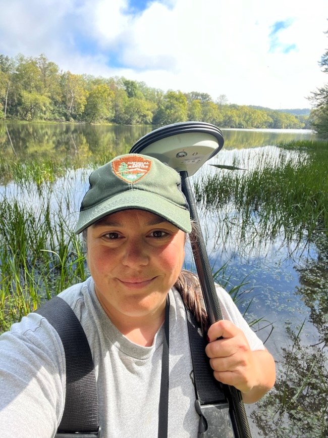 Layne Strickler Smiling park ranger holding a GPS surveying device, standing in front of a lake surrounded by tall green grass